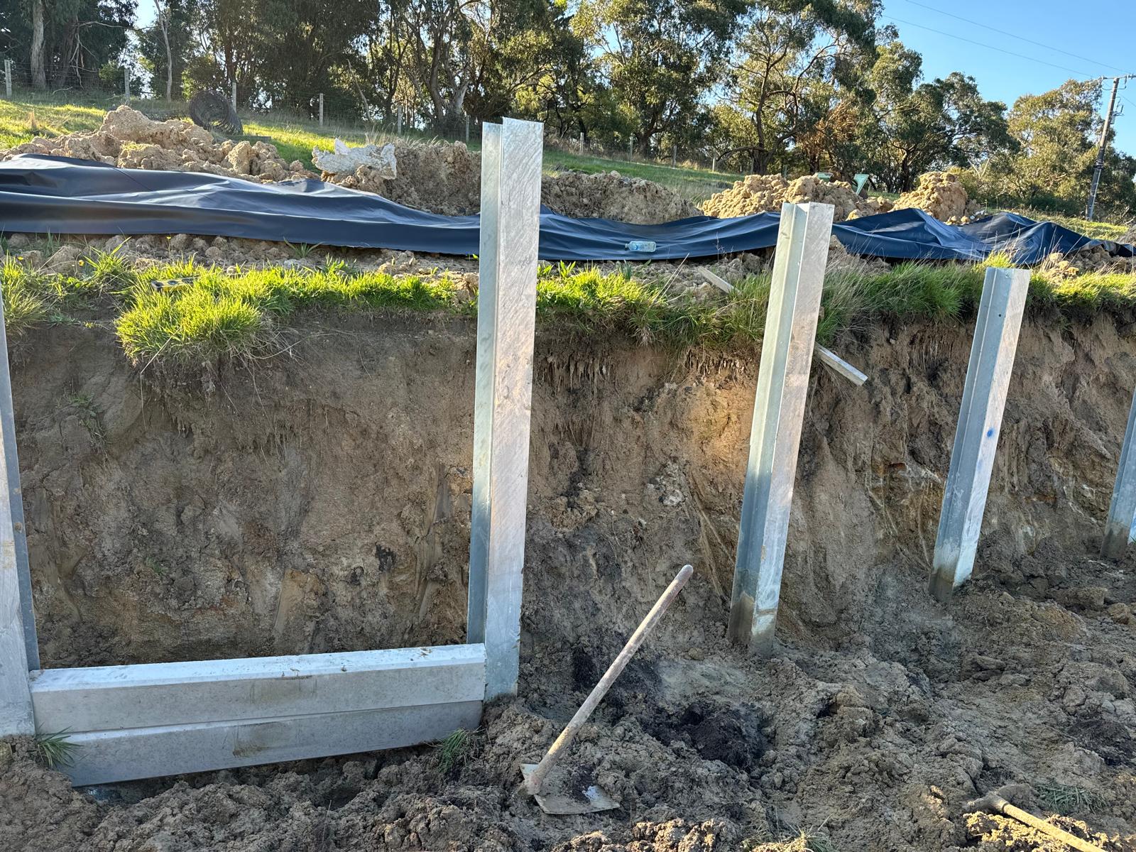 Steel retaining wall posts set into a cut bank with the first concrete sleepers in place