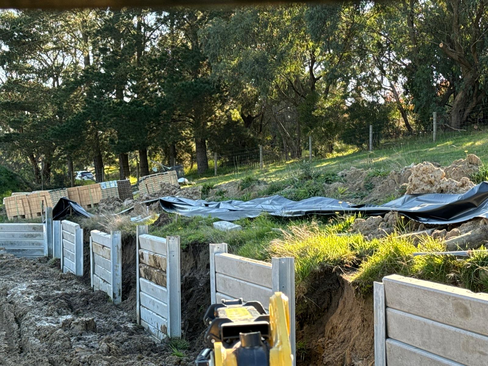 A concrete sleeper retaining wall going in along a sloping bank, with a laser level in the foreground